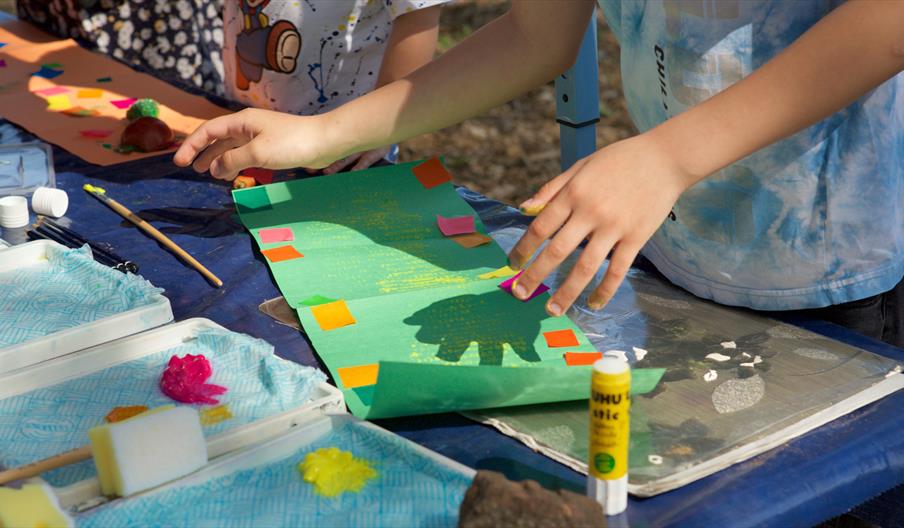 An image of a child sticking colored pieces of paper onto a green piece of paper, surrounded by a glue stick and a paintbrush.