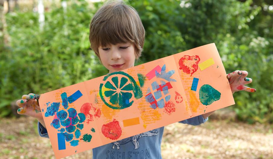 An image of a child proudly holding up a piece of their stamped artwork. The stamps include images of fruits and other block shapes