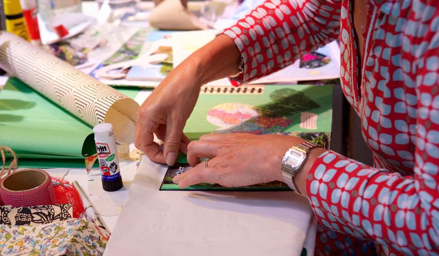 An image of a woman wearing a red and blue long sleeve shirt sticking items down onto a collage of colored paper