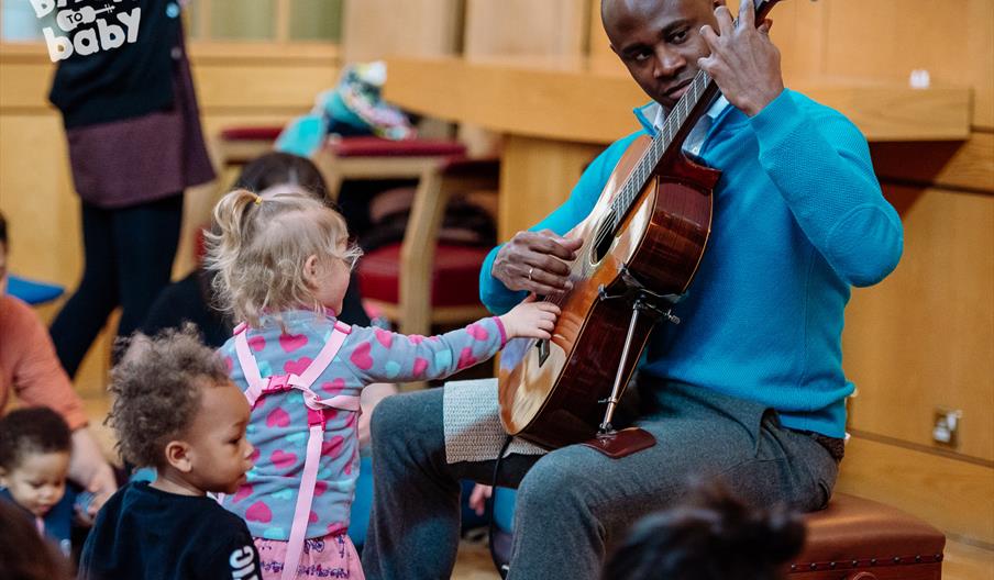 A man playing the guitar. A toddler is standing in front of him and copying him, reaching out to touch the strings