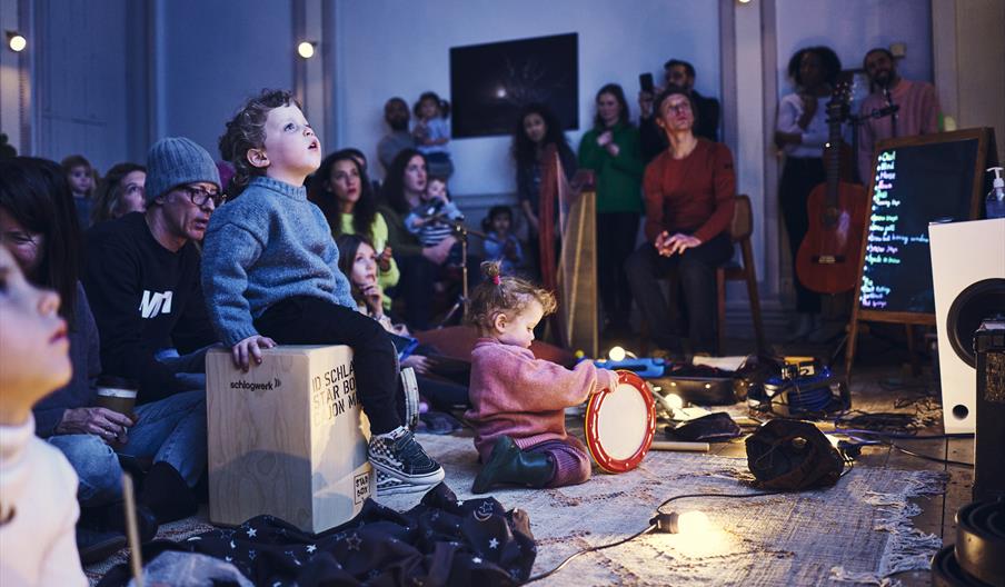 An image from an Enchanted Cinema show. In the centre of the image is young person sitting on a wooden block looking up at a projection. This person i