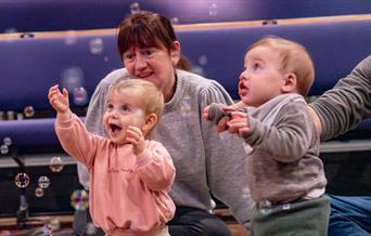 A baby in a pink jumper looks up in awe at a colourful toy held above their head by a woman wearing a patterned shirt and jeans. She kneels on the flo