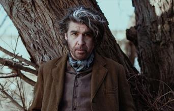 A man is stood in front of a wildly defined tree. He has wavy, black and grey hair with a deep side part. He is wearing a chocolate brown waistcoat, a