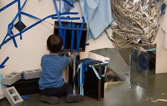 A child plays with blue tape and silver materials in a gallery.