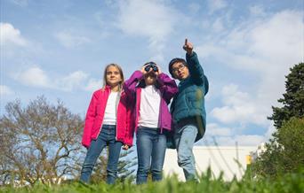 Three children, one of them pointing at something and the other looking at it with a pair of binoculars