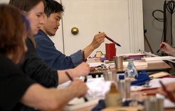 A group of artists , sitting at tables and holding mop-brushes, focus on their artworks at an Artanda workshop.