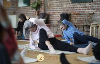 Two women sitting on the floor are gently following an Awareness Through Movement Lesson: leaning on their right elbow, the other hand placed on the h