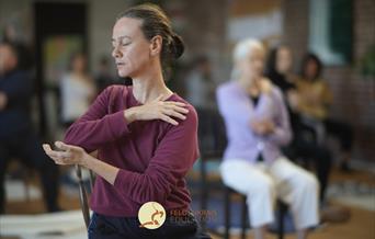 A women is gently moving following the instruction of the Awareness Through Movement guided by the teacher. she has her eyes closed, sitting on a chai