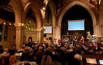 An audience sits at round tables in a festively lit church. At the front, musicians are playing a grand piano and a clarinet.