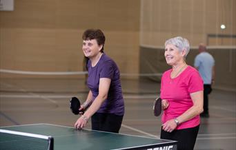 An image of two women with Parkinson's taking part in a friendly table tennis match together. One woman is wearing a purple top with black trousers an