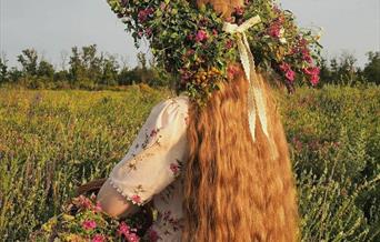 woman with the wreath on her head standing with her back to us in the summer field