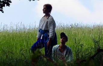 A standing woman and a seated man both with pale skin occupy the shady long grass beneath an oak tree. In the background is the brow of a grassy hill