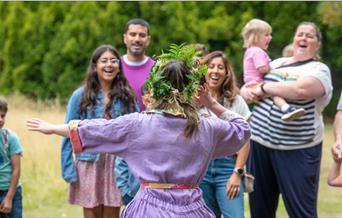 A woman in a purple jumpsuit wearing a crown made of leaves, stands with her back to the camera, gestures to laughing families