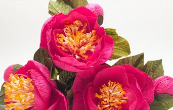 Three vibrant pink flowers crafted out of crepe paper assembled in a mini-bouquet in front of a studio backdrop