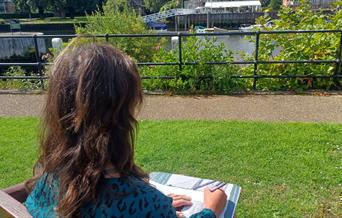 Image of a woman with long brown hair sat sketching near the Thames, taken from over her shoulder