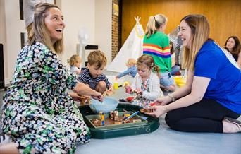 A parent and facilitator at a Ways To Play Today class, with children playing in a tray of animals and taste-safe mud