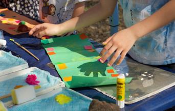 An image of a child sticking colored pieces of paper onto a green piece of paper, surrounded by a glue stick and a paintbrush.