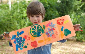 An image of a child proudly holding up a piece of their stamped artwork. The stamps include images of fruits and other block shapes
