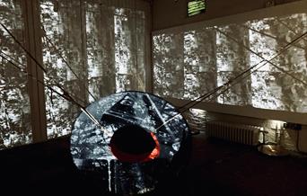 A large fabric sculpture in the shape of a ring, with a projection on it. Abstract projections of cylinders are behind