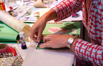 An image of a woman wearing a red and blue long sleeve shirt sticking items down onto a collage of colored paper