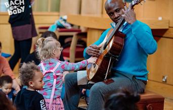 A man playing the guitar. A toddler is standing in front of him and copying him, reaching out to touch the strings