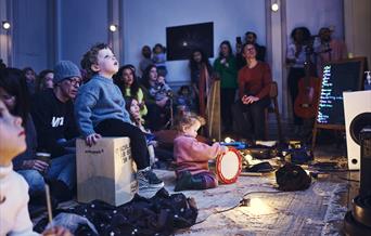 An image from an Enchanted Cinema show. In the centre of the image is young person sitting on a wooden block looking up at a projection. This person i