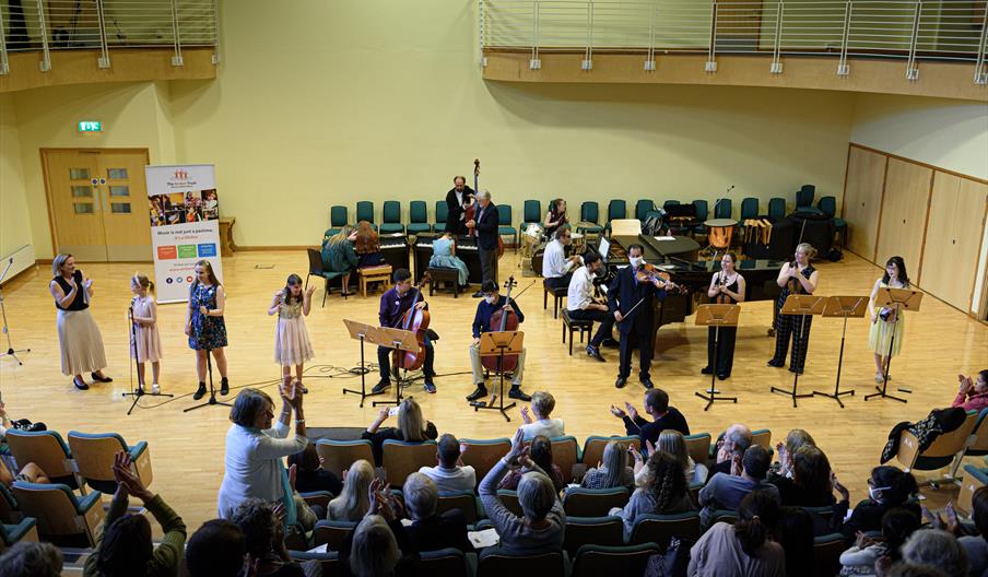 Musicians including singers, cellists, violinists, and pianists are performing together at an indoor stage in front of an audience who applauds.