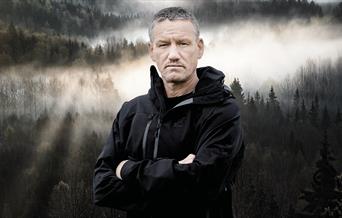 Mark Billingham stands facing the camera with his arms crossed and a serious expression on his face. He is against a backdrop of a misty mountain fore