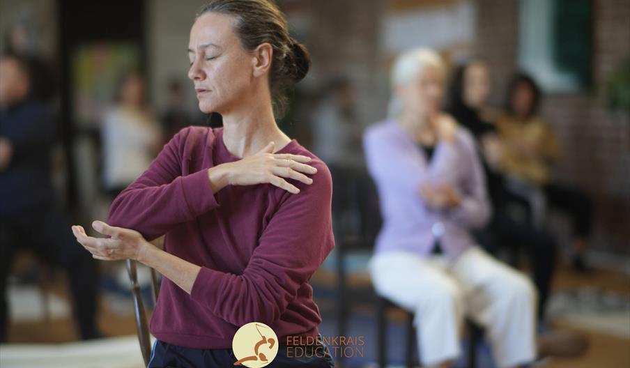 A women is gently moving following the instruction of the Awareness Through Movement guided by the teacher. she has her eyes closed, sitting on a chai