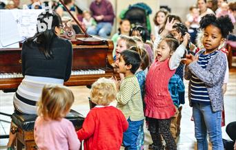 A pianist in a stripy top playing a piano with her back to the camera. A small crowd of happy children with their hands in the air around her