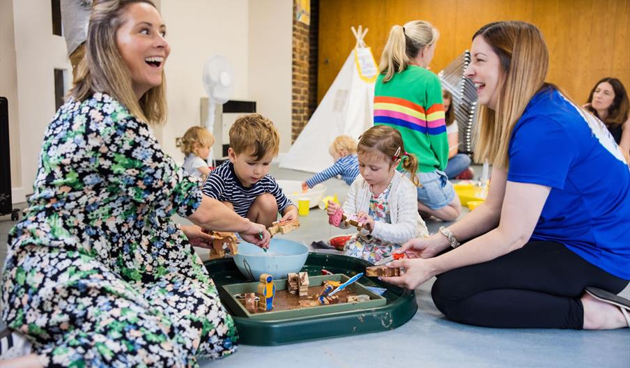 A parent and facilitator at a Ways To Play Today class, with children playing in a tray of animals and taste-safe mud A parent and facilitator at a Ways To Play Today class, with children playing in a tray of animals and taste-safe mud