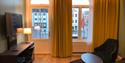 Nyoppusset rom Thon Hotel Høyers med gule gardiner som går fra gulv til tak, 2 buende vinduer, en stol og et ovalt bord, skrivepult og et tv.