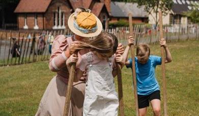 A girl and a boy on stilts being helped by a lady in Victorian costume.