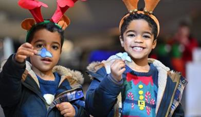 two young children wearing reindeer antlers and Christmas jumpers under their coats.