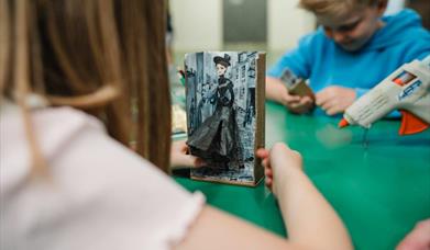 Girl playing with a mechanical toy that she has made to look like a Victorian doll, there is a boy in the background and an adult holding a glue gun t