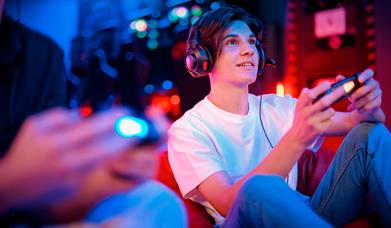 Teenage boy smiling and playing on a game, holding a controller, sat next to his friend.