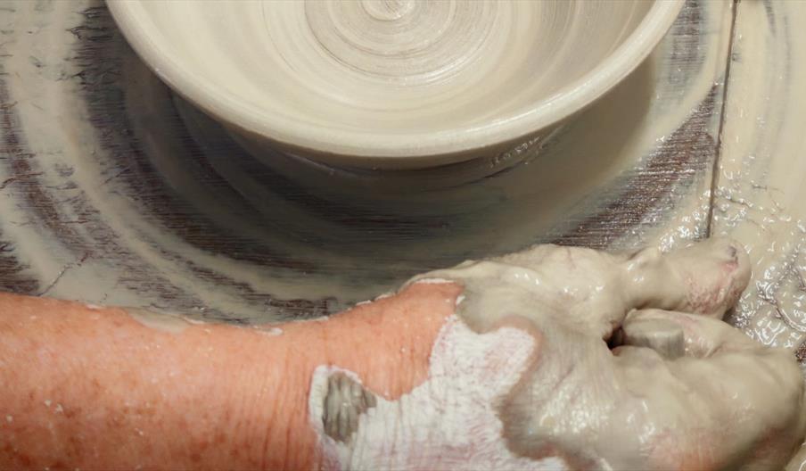 A close-up of hands shaping a wide clay bowl on a spinning potter's wheel. The bowl is smooth and centred, with spiral lines visible inside. Wet clay
