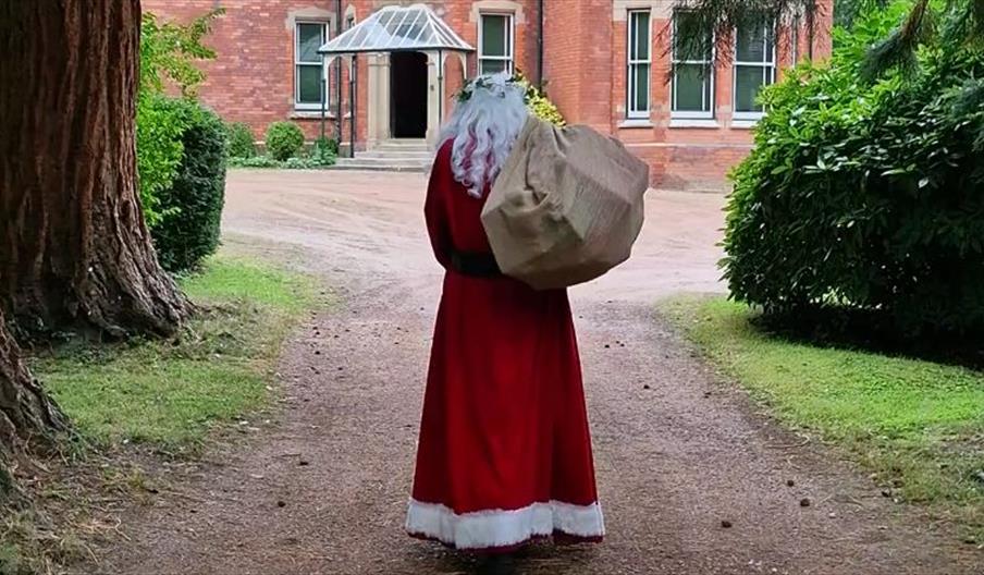 Father Christmas, dressed in his iconic red suit with white trim, walks down a tree-lined path toward a large, red-brick Victorian-style house. He is