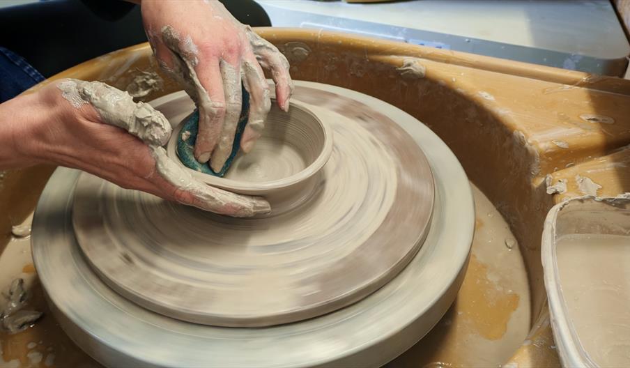 Several people learning pottery on wheels in a studio, with clay-covered hands shaping bowls as the wheels spin. The foreground shows a close-up of ha