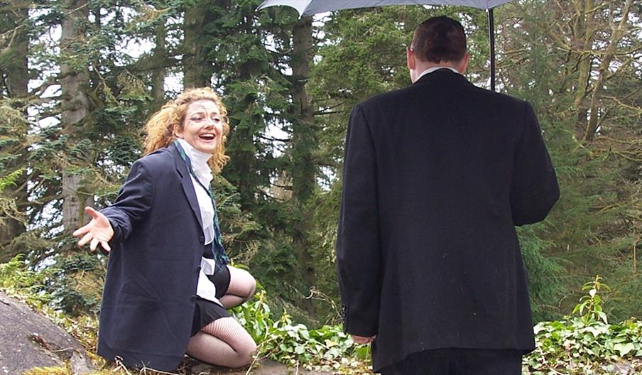 Two people meeting in a woodland setting; a woman crouching on a wall and a man holding an umbrella over his head.