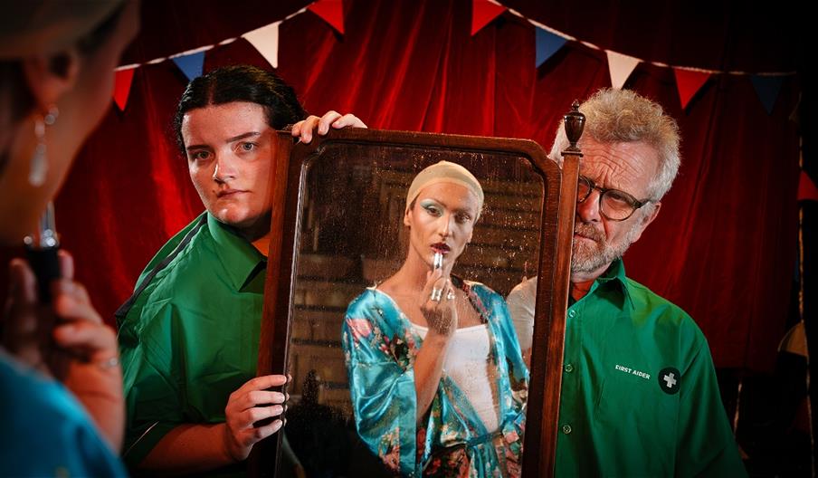 A man and a woman wearing green medic uniforms hold a mirror in which is the reflection of a man wearing a silky blue robe and applying make-up.