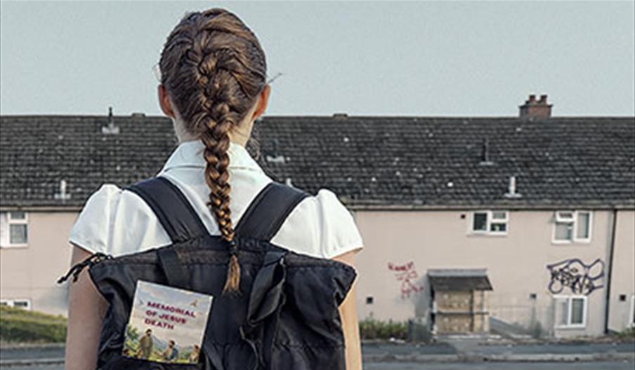 A schoolgirl with a long plait faces away from the camera towards a row of houses which have graffiti on them. The girl is wearing a backpack with a b