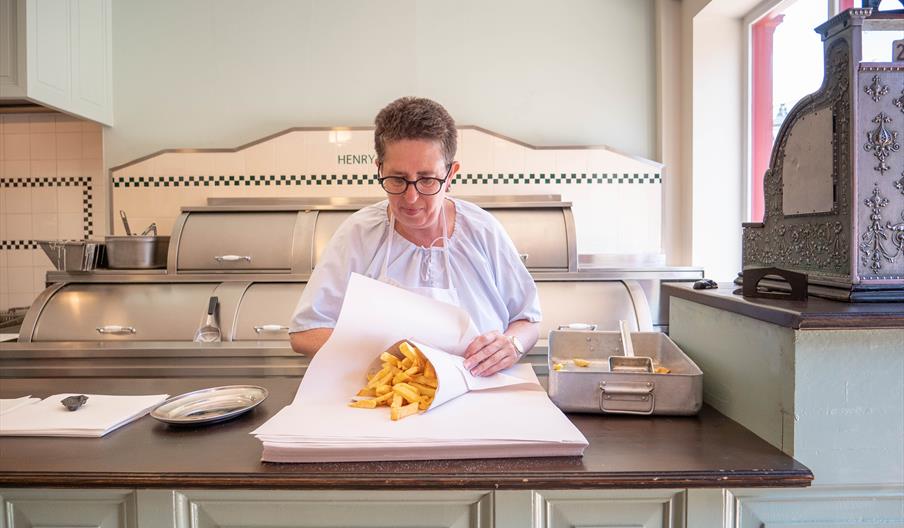 A lady wrapping traditionally cooked chips in paper in a Victorian chip shop at Blists HIll Victorian Town