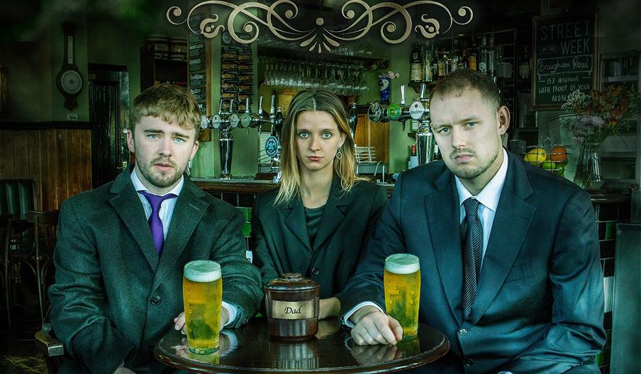 Two men and a woman wearing dark clothes sit at a pub table. Each man has a pint of lager. In front of the woman is a cremation urn labelled 'Dad'.