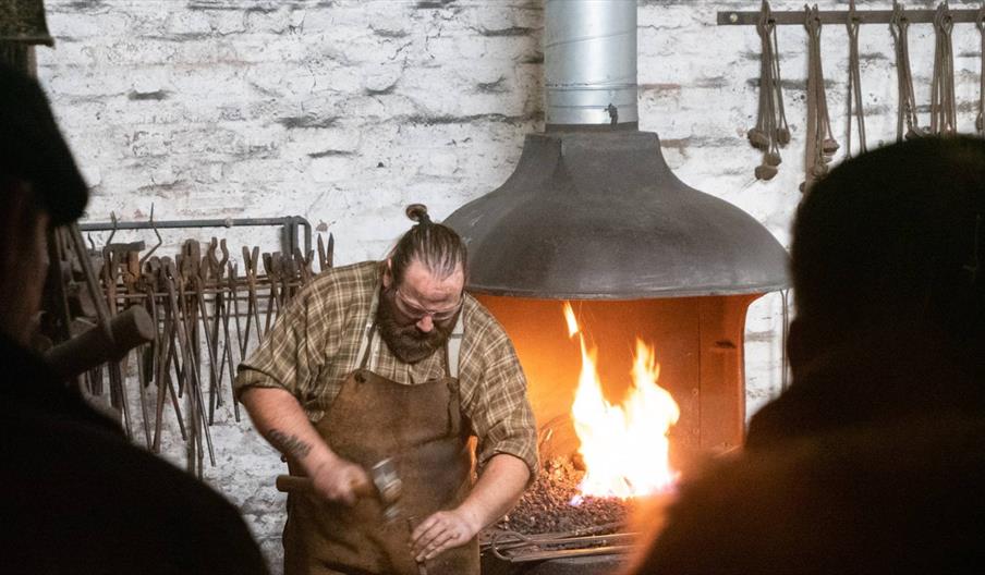 The image shows a blacksmith wearing a leather apron, in front of the forge.