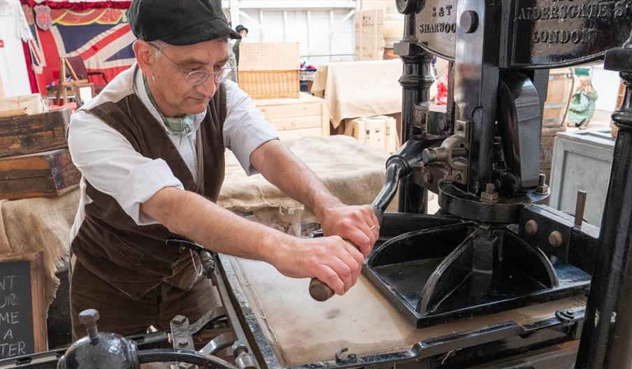 A man dressed in Victorian clothing is using a manual printing press