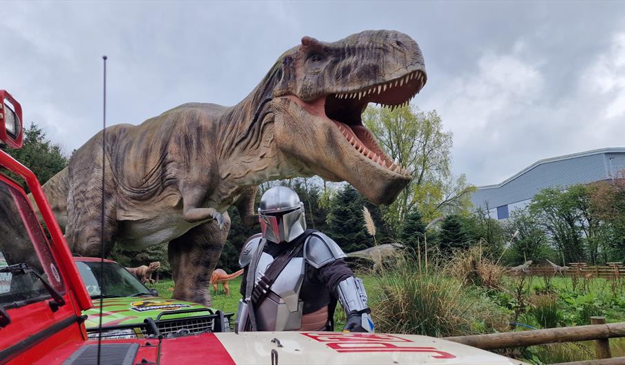 The Mandalorian standing by a vehicle with a Hoo Zoo T-Rex behind.