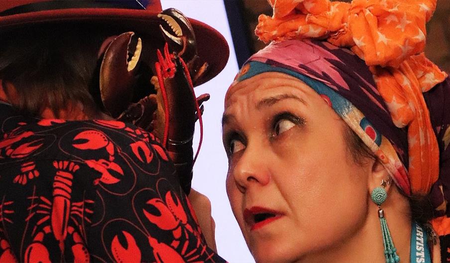 Agent Kahlo, wearing large blue earrings and a flamboyant headscarf, leans in to whisper to Agent Dali who is wearing a coat patterned with lobsters.