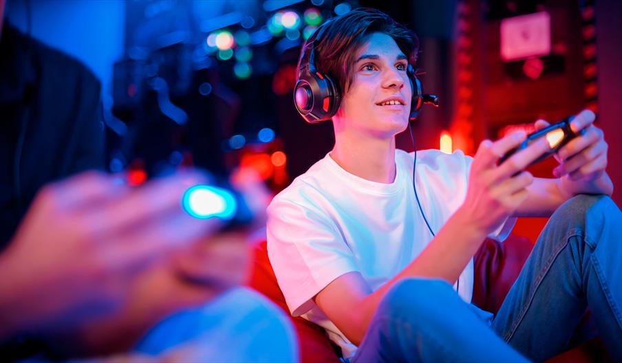 Teenage boy smiling and playing on a game, holding a controller, sat next to his friend.