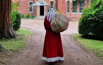 Father Christmas, dressed in his iconic red suit with white trim, walks down a tree-lined path toward a large, red-brick Victorian-style house. He is